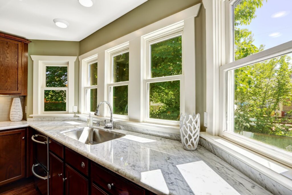Bright kitchen with marble countertops, sink, and large windows overlooking greenery.