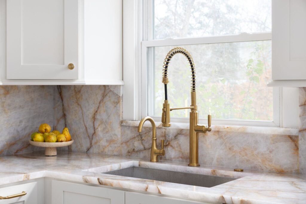 Elegant kitchen sink area featuring a gold pull-down faucet, marble countertops and backsplash, and white cabinetry.